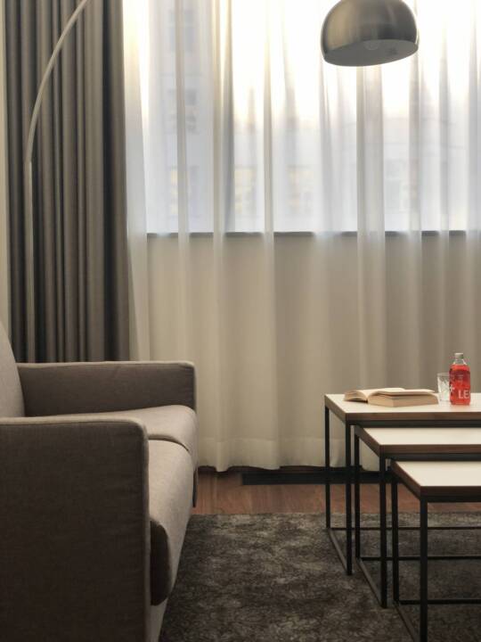 Bright hotel room at the MOA Berlin in Moabit with a grey sofa, two nested coffee tables, an open book and a red drinks bottle. Light-coloured curtains hang in front of the window, next to which stands a tall, curved floor lamp.