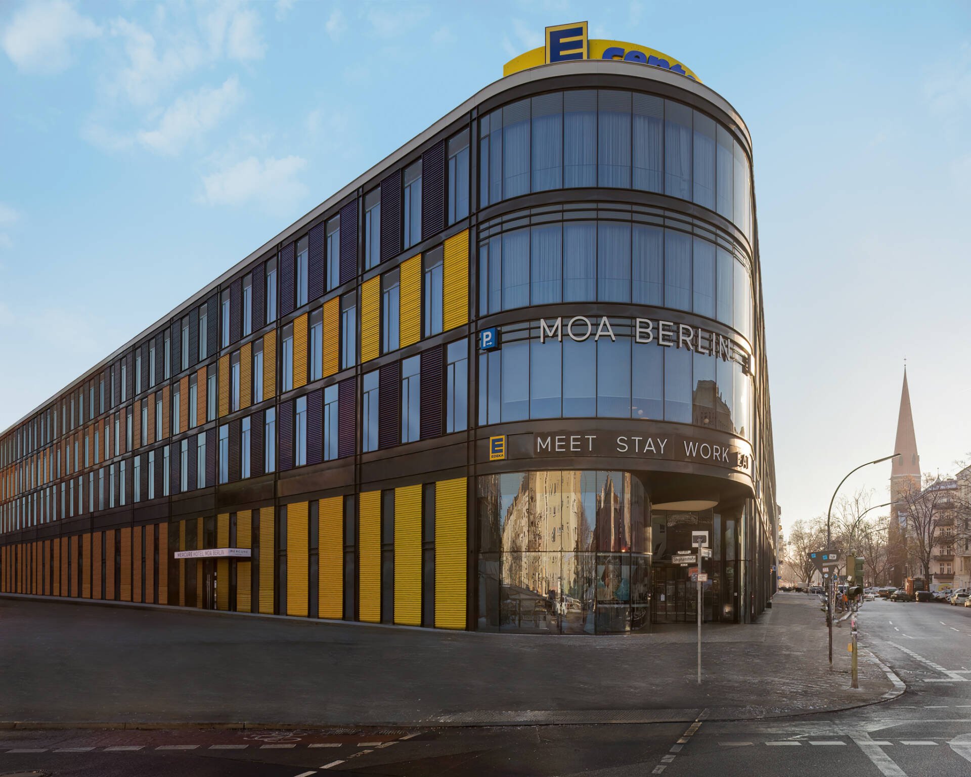 Die Außenansicht des Hotels MOA Berlin in Moabit: ein modernes, mehrstöckiges Gebäude mit abgerundeter Glasfront, gelben und braunen Fassadenelementen und dem Schriftzug "MOA BERLIN".