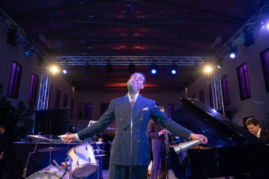 A man in a suit stands with his arms outstretched in front of a drum kit and grand piano on a floodlit stage in Hotel MOA Berlin in Mitte.